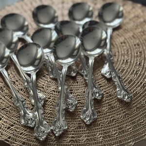 May include: A collection of ornate silver soup spoons arranged on a decorative, woven placemat. The spoons have a reflective surface and detailed floral patterns on the handles. The spoons are a classic design.