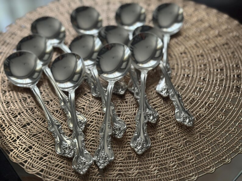 May include: A collection of ornate silver soup spoons arranged on a decorative, woven placemat. The spoons have a reflective surface and detailed floral patterns on the handles. The spoons are a classic design.