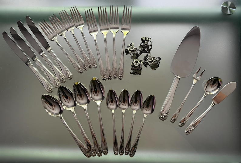 Pu&ograve; includere: Un set di posate color argento che include coltelli, forchette e cucchiai, insieme a una paletta per torte, una piccola forchetta a due rebbi e quattro portatovaglioli in metallo decorativi con motivo floreale su una superficie riflettente.
