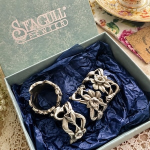 Pu&ograve; includere: Un set di tre portatovaglioli in peltro color argento con un design floreale in rilievo a forma di narciso. Gli articoli sono presentati all'interno di una scatola azzurra del marchio Seagull Pewter foderata con carta velina blu scuro.