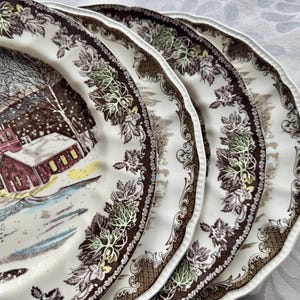 May include: Three brown and white ceramic dinner plates with a winter scene design. The plates feature a brown and white floral border with a scalloped edge.