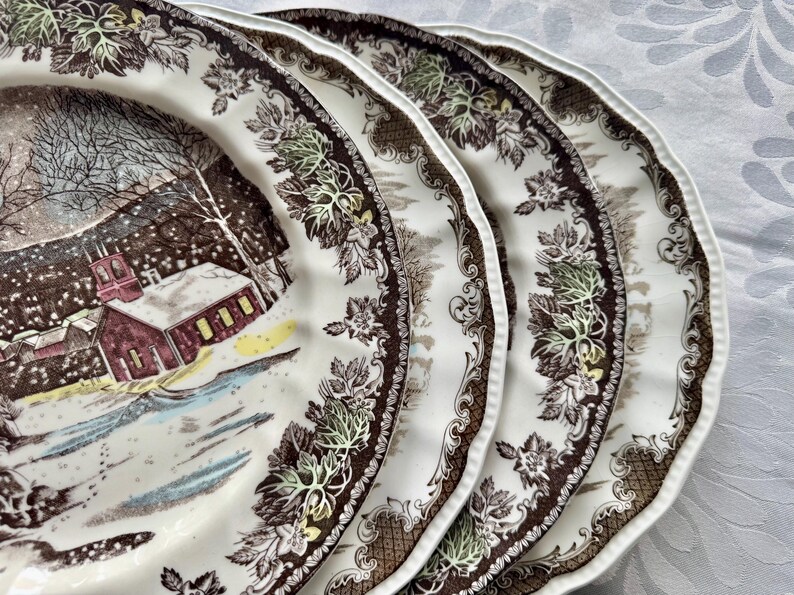 May include: Three brown and white ceramic dinner plates with a winter scene design. The plates feature a brown and white floral border with a scalloped edge.