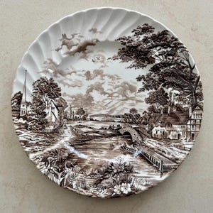 May include: A round white ceramic plate with a scalloped edge featuring a detailed brown transferware print of a rural landscape. The scene depicts a river, a stone bridge, a small boat, houses, a church spire, and trees under a cloudy sky.