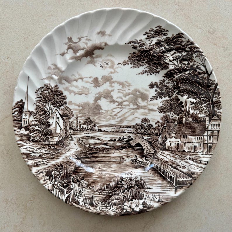 May include: A round white ceramic plate with a scalloped edge featuring a detailed brown transferware print of a rural landscape. The scene depicts a river, a stone bridge, a small boat, houses, a church spire, and trees under a cloudy sky.