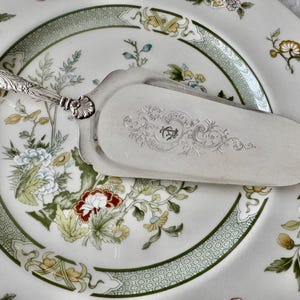 K&ouml;nnte beinhalten: Ein silberner Tortenheber mit einem verzierten Griff liegt auf einem Teller mit floralem Muster. Der Teller hat einen gr&uuml;nen Rand und ein Design aus Blumen in Rot, Blau und Gelb. Die Klinge des Hebers hat ein eingraviertes Design.