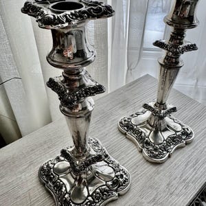 May include: A pair of silver-toned metal candlesticks featuring ornate, embossed floral and scrollwork detailing on the square bases, the central stems, and the top candle holders. The metallic surface has a polished, reflective finish.