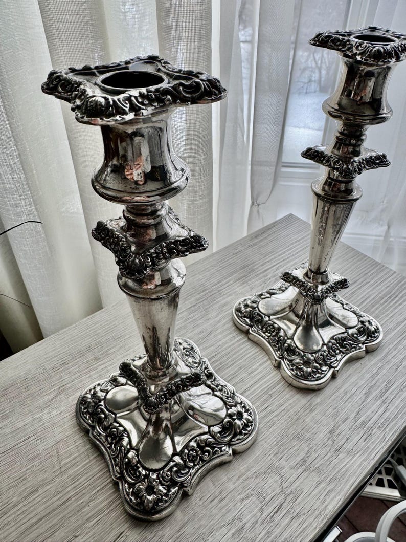May include: A pair of silver-toned metal candlesticks featuring ornate, embossed floral and scrollwork detailing on the square bases, the central stems, and the top candle holders. The metallic surface has a polished, reflective finish.