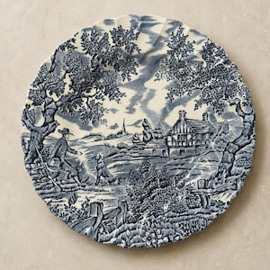 May include: A round ceramic plate with a scalloped edge featuring a blue and white transferware pattern. The design depicts a rural landscape with a person holding a rifle, a dog, a timber-framed house, large trees, and a distant church spire.