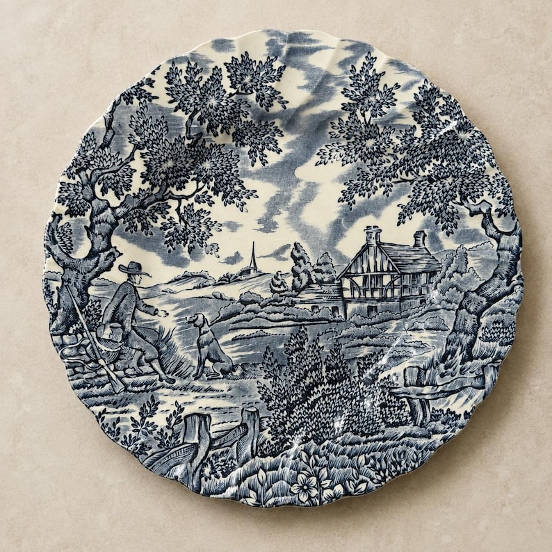 May include: A round ceramic plate with a scalloped edge featuring a blue and white transferware pattern. The design depicts a rural landscape with a person holding a rifle, a dog, a timber-framed house, large trees, and a distant church spire.