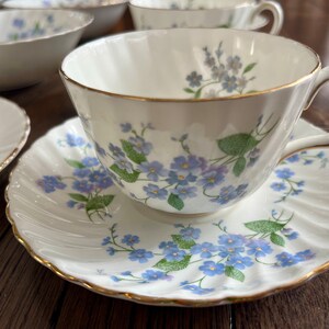 May include: A white teacup and saucer set with a delicate blue floral pattern and gold trim. The teacup has a curved handle and sits on a matching saucer. Other matching bowls and plates are visible in the background.