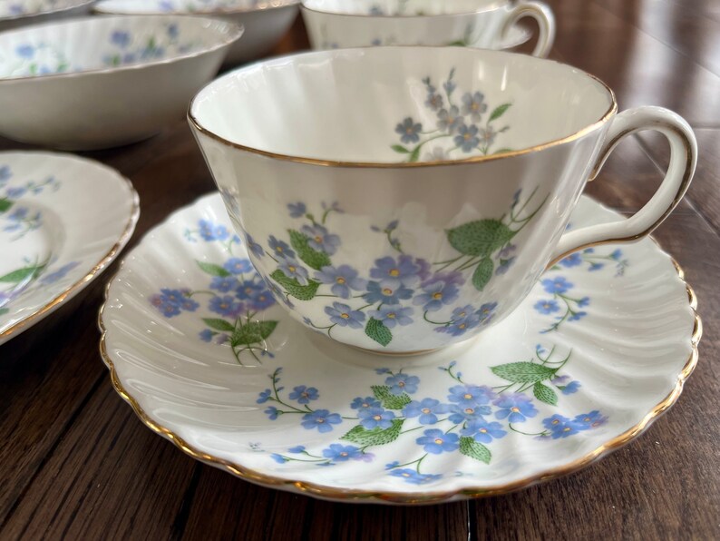 May include: A white teacup and saucer set with a delicate blue floral pattern and gold trim. The teacup has a curved handle and sits on a matching saucer. Other matching bowls and plates are visible in the background.
