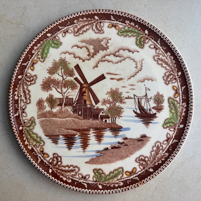 May include: A round ceramic plate featuring a brown and cream transferware scene of a windmill near a body of water with a sailboat. The border is decorated with brown oak leaves and acorns, with small accents of green leaves throughout the design.