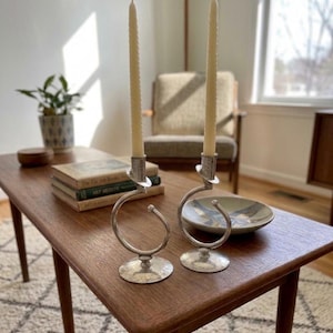 Peut inclure: Deux bougeoirs argent&eacute;s avec des motifs en spirale, chacun tenant une longue bougie cr&egrave;me. Les bougeoirs sont pos&eacute;s sur une table en bois avec des livres et un bol d&eacute;coratif. La pi&egrave;ce est bien &eacute;clair&eacute;e.