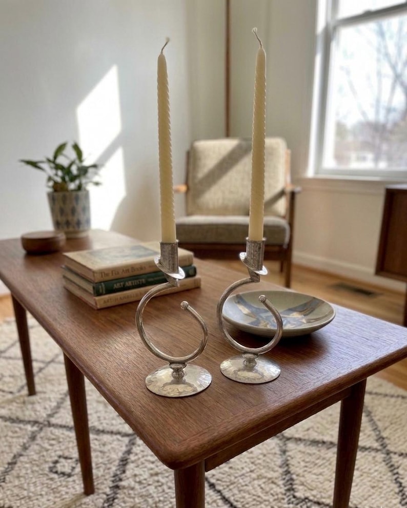 Peut inclure: Deux bougeoirs argent&eacute;s avec des motifs en spirale, chacun tenant une longue bougie cr&egrave;me. Les bougeoirs sont pos&eacute;s sur une table en bois avec des livres et un bol d&eacute;coratif. La pi&egrave;ce est bien &eacute;clair&eacute;e.