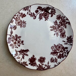 May include: A round white ceramic plate with a scalloped edge, featuring a dark brown transferware pattern of blooming flowers, leafy branches, and small birds perched among the foliage, arranged in a circular border design.