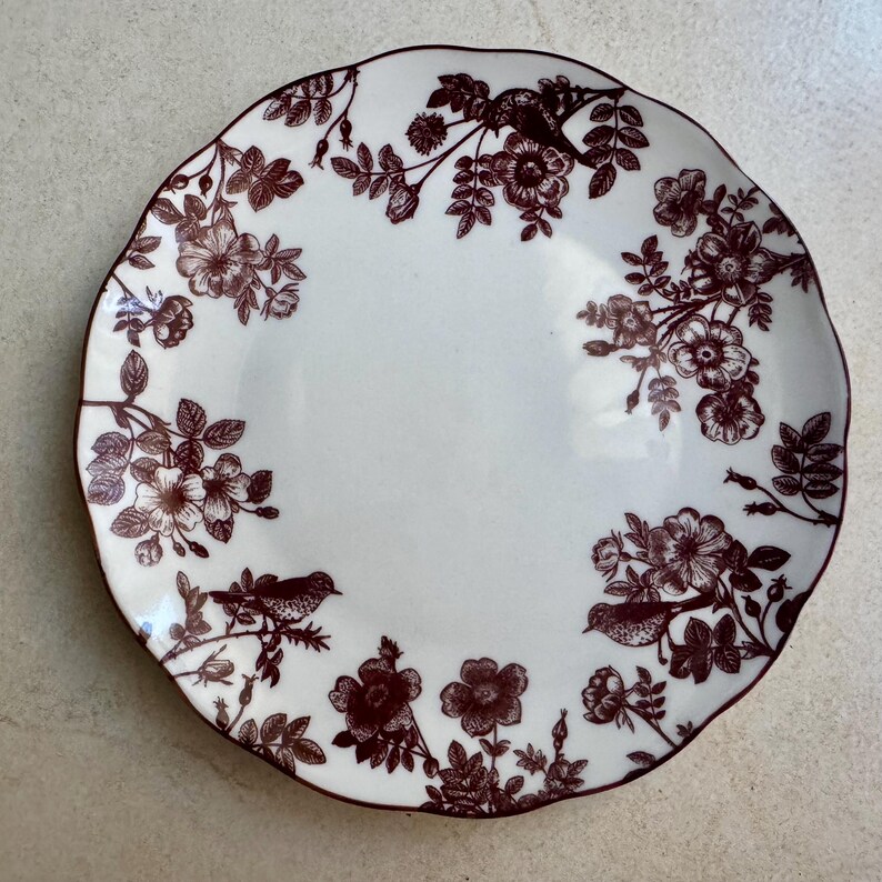 May include: A round white ceramic plate with a scalloped edge, featuring a dark brown transferware pattern of blooming flowers, leafy branches, and small birds perched among the foliage, arranged in a circular border design.
