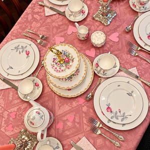 May include: A tea party table setting featuring white porcelain plates, cups, and saucers decorated with pink rose motifs, a tiered serving stand, silver-toned cutlery, two ornate candle holders with coral candles, and pink paper hearts.