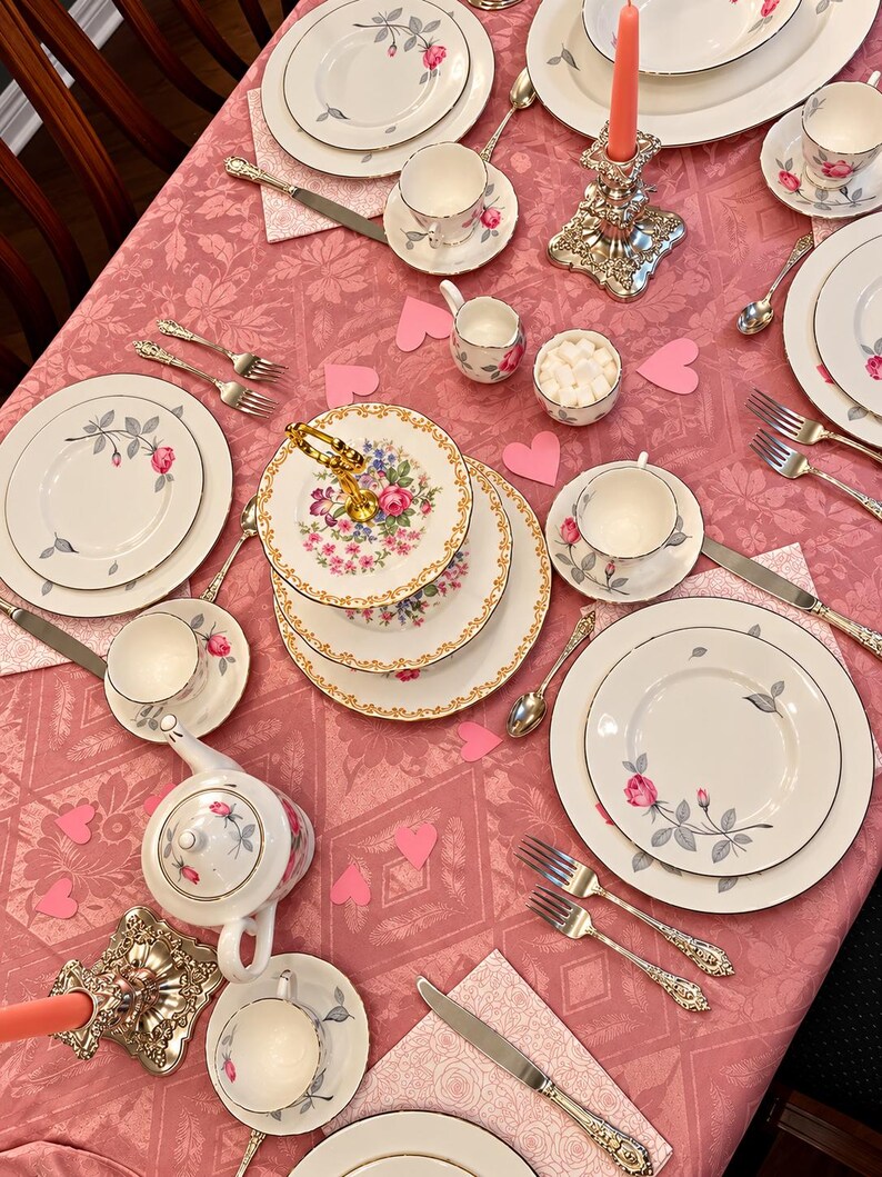 May include: A tea party table setting featuring white porcelain plates, cups, and saucers decorated with pink rose motifs, a tiered serving stand, silver-toned cutlery, two ornate candle holders with coral candles, and pink paper hearts.