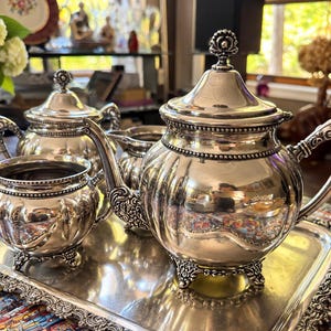 May include: A silver tea set on a rectangular silver tray, including a teapot with a lid, a lidded sugar bowl, a creamer, and an open sugar bowl. Each piece features a polished finish, ornate scrollwork handles, and decorative feet.