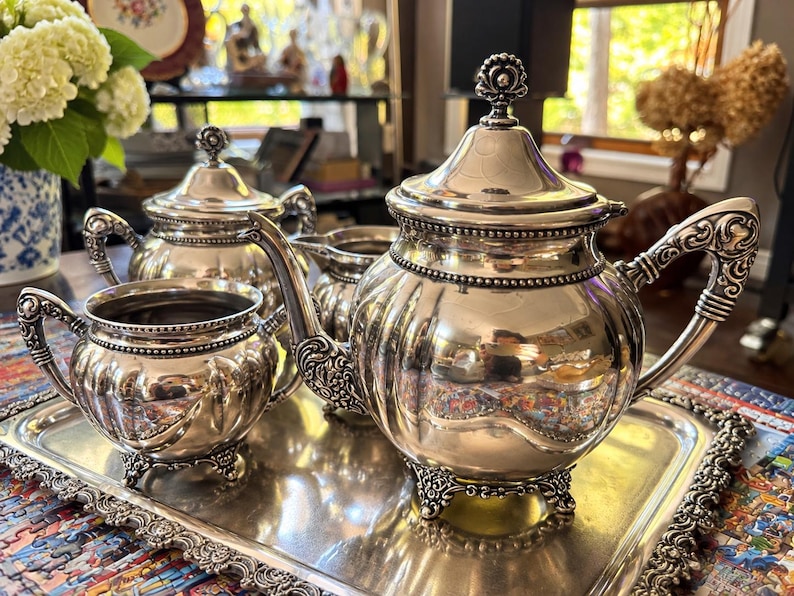 May include: A silver tea set on a rectangular silver tray, including a teapot with a lid, a lidded sugar bowl, a creamer, and an open sugar bowl. Each piece features a polished finish, ornate scrollwork handles, and decorative feet.