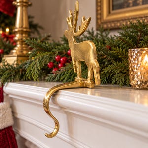 May include: A gold-colored metal stocking holder shaped like a standing reindeer with large antlers. The figure is attached to a curved hook designed to hang over the edge of a mantelpiece, featuring a smooth, reflective metallic finish.