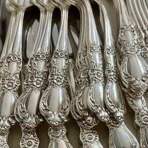 May include: A collection of silver-toned metal flatware handles arranged in a row, each featuring intricate embossed floral and scrollwork patterns along the stems and rounded ends, with a polished, reflective metallic finish.