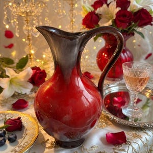 May include: A red and brown ceramic pitcher with a handle, set on a table with a floral arrangement and a glass of wine. The pitcher has a glossy finish and a rounded body. A plate with blueberries and a rose is also visible.