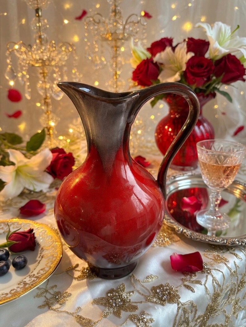 May include: A red and brown ceramic pitcher with a handle, set on a table with a floral arrangement and a glass of wine. The pitcher has a glossy finish and a rounded body. A plate with blueberries and a rose is also visible.