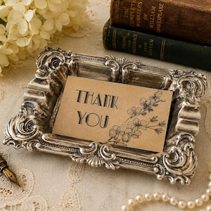 May include: A rectangular silver-toned metal tray with ornate, raised scrollwork edges. Inside the tray sits a rectangular tan card featuring the black printed text THANK YOU and a detailed black line drawing of a flowering branch.