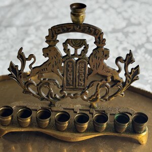 May include: A brass menorah with eight candle holders and a central shamash holder. The decorative piece features two lions flanking a central panel with Hebrew text. The menorah sits on a brass tray, with a white patterned background.