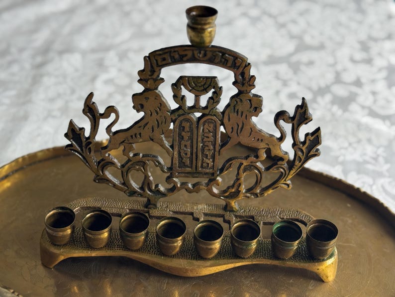 May include: A brass menorah with eight candle holders and a central shamash holder. The decorative piece features two lions flanking a central panel with Hebrew text. The menorah sits on a brass tray, with a white patterned background.