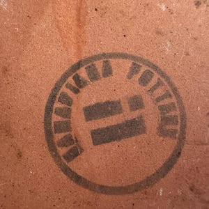 May include: Close-up of a textured, reddish-brown surface with a circular black stamp. The stamp reads "KAMA POTTERY" in a circular pattern around a central design.