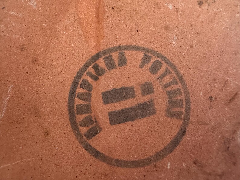 May include: Close-up of a textured, reddish-brown surface with a circular black stamp. The stamp reads "KAMA POTTERY" in a circular pattern around a central design.