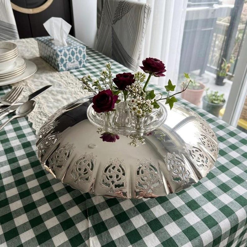 May include: A silver-colored, decorative serving dish with an ornate, scalloped edge. A small glass vase holds dark red roses and greenery. The dish sits on a green and white checkered tablecloth, with a lace runner and silverware visible.
