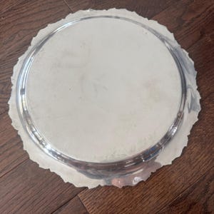 May include: A round silver-toned metal serving tray with a smooth, reflective center and a decorative, scalloped edge. The tray features a polished finish and a subtle, embossed hallmark stamp visible near the upper rim of the circular surface.