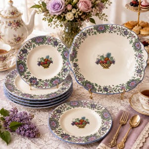 May include: A set of cream-colored ceramic dinnerware featuring a decorative border of purple and blue flowers with green vines. Each piece displays a central illustration of a woven basket filled with colorful fruit, including grapes and pineapple.