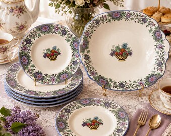 May include: A set of cream-colored ceramic dinnerware featuring a decorative border of purple and blue flowers with green vines. Each piece displays a central illustration of a woven basket filled with colorful fruit, including grapes and pineapple.