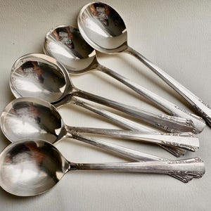 May include: Five silver soup spoons with ornate handles. The spoons are arranged in a fan shape, with the handles pointing towards the top of the image.