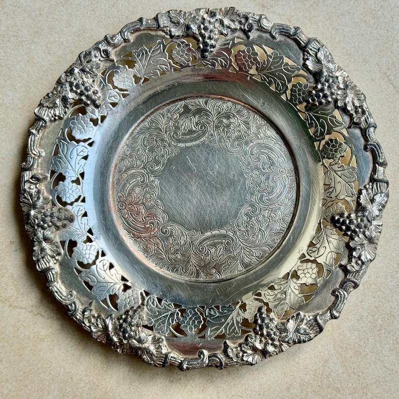 May include: A round silver-toned metal tray featuring an intricate openwork border decorated with embossed grape clusters and vine leaves. The central area of the tray displays a detailed engraved pattern with swirling floral motifs.