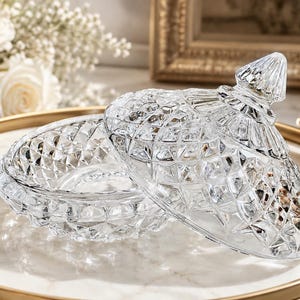 May include: A clear glass candy dish with a matching lid, featuring a faceted diamond-cut pattern throughout the exterior. The bowl has a circular base, and the lid is topped with a pointed, textured finial, creating a reflective surface.