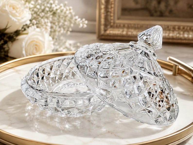 May include: A clear glass candy dish with a matching lid, featuring a faceted diamond-cut pattern throughout the exterior. The bowl has a circular base, and the lid is topped with a pointed, textured finial, creating a reflective surface.