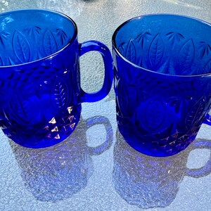 May include: Two cobalt blue glass mugs with a leaf pattern and a diamond-shaped design. The mugs have handles.