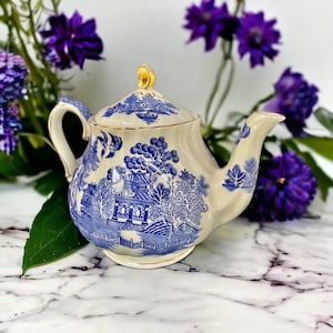 May include: Cream-coloured teapot with a blue Willow pattern design. The teapot has a curved spout, a handle, and a lid with a gold finial. The design features a landscape scene with buildings and trees. A classic piece for tea lovers.