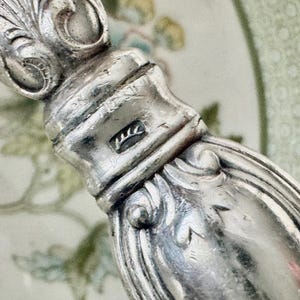 K&ouml;nnte beinhalten: Nahaufnahme eines silbernen Besteckgriffs mit aufwendigen Schn&ouml;rkeln und einem Stempel. Der Griff hat eine polierte, reflektierende Oberfl&auml;che. Der Hintergrund ist ein verschwommenes Blumenmuster.