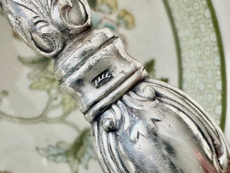 K&ouml;nnte beinhalten: Nahaufnahme eines silbernen Besteckgriffs mit aufwendigen Schn&ouml;rkeln und einem Stempel. Der Griff hat eine polierte, reflektierende Oberfl&auml;che. Der Hintergrund ist ein verschwommenes Blumenmuster.