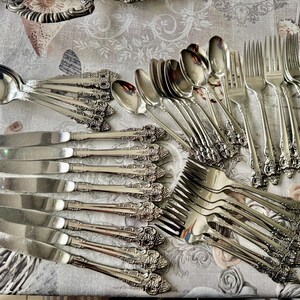 May include: An assortment of vintage silverware, including spoons, knives, and forks, arranged on a floral patterned tablecloth. The silverware has ornate, detailed handles and a shiny, reflective surface. The arrangement suggests a formal dining setting.