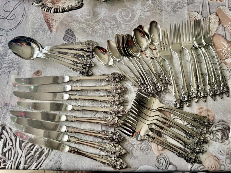 May include: An assortment of vintage silverware, including spoons, knives, and forks, arranged on a floral patterned tablecloth. The silverware has ornate, detailed handles and a shiny, reflective surface. The arrangement suggests a formal dining setting.