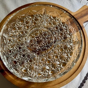 May include: A clear glass serving platter with a starburst pattern, set on a round wooden tray with a handle. The platter is divided into sections, perfect for serving snacks or appetizers. The wooden tray has a diameter of approximately 12 inches.