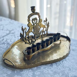 May include: A brass menorah with a decorative lion and shield design, set on a matching oval tray. The menorah features eight candle holders and a central shamash holder. The tray has handles and a textured surface.