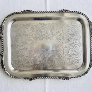 Pu&ograve; includere: Un vassoio da portata rettangolare color argento caratterizzato da un motivo a volute inciso sulla superficie centrale. Il vassoio include due manici decorativi sui lati e un bordo rialzato con un dettaglio ripetitivo a forma di conchiglia e corda.